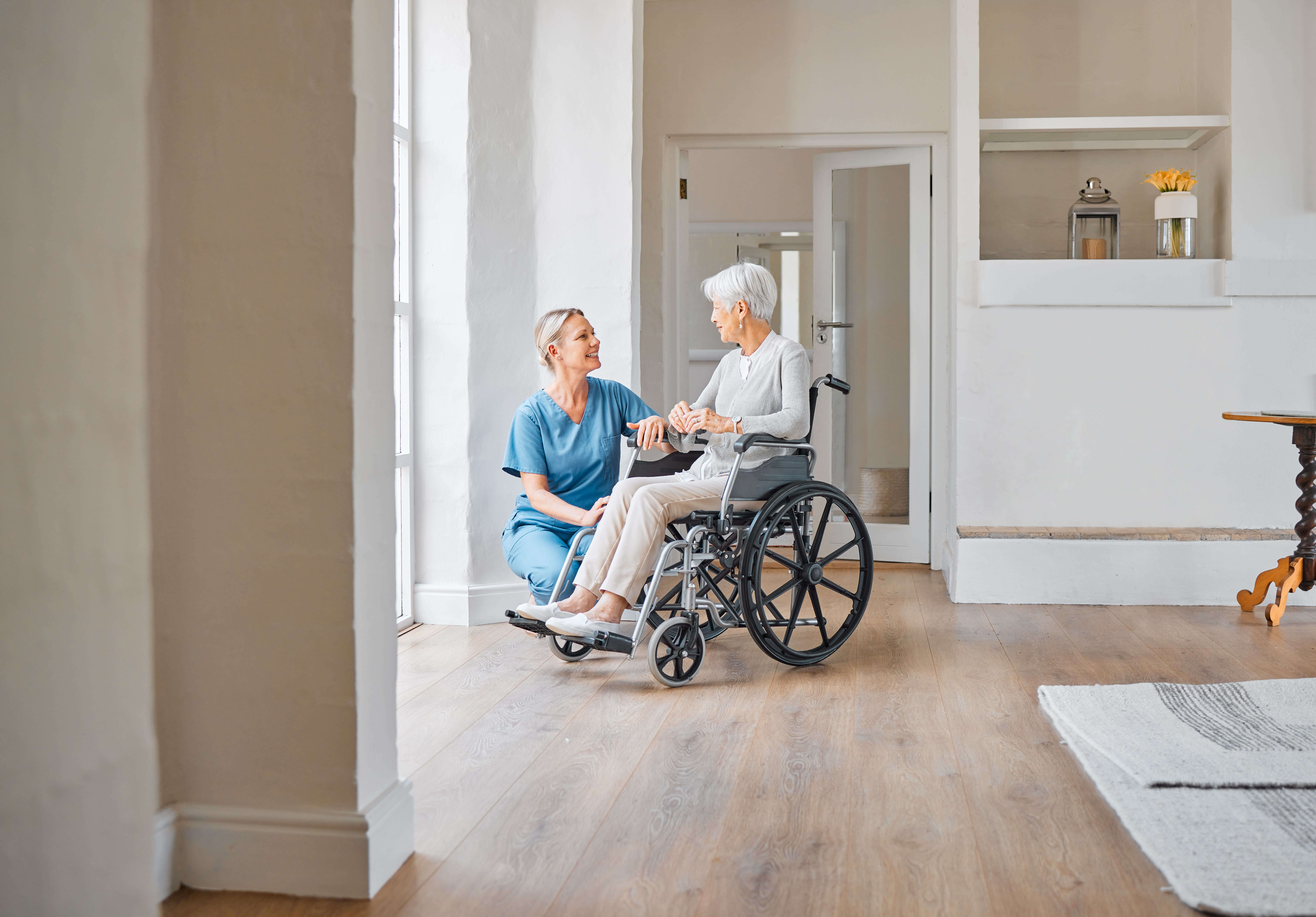 Pflegekraft im Gespräch mit älterer Frau im Rollstuhl, helle Wohnumgebung